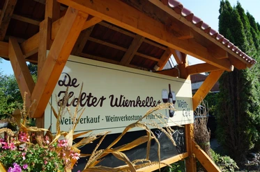 Weincafé De Holter Wienkeller Schild mit der Aufschrift De Holter Wienkeller und Angeboten wie Weinverkauf, Weinverkostung und Frühstück unter einem Holzdach
