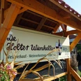 Weincafé De Holter Wienkeller Schild mit der Aufschrift De Holter Wienkeller und Angeboten wie Weinverkauf, Weinverkostung und Frühstück unter einem Holzdach