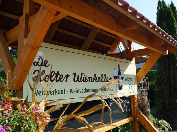 Weincafé De Holter Wienkeller Schild mit der Aufschrift De Holter Wienkeller und Angeboten wie Weinverkauf, Weinverkostung und Frühstück unter einem Holzdach