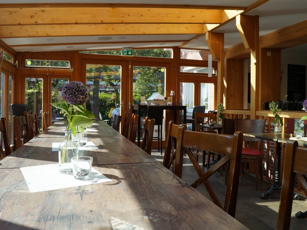 Weincafé De Holter Wienkeller, Innenansicht, Gemeinde Rhauderfehn Langer Holztisch in einem hellen Weincafé mit Holzstühlen und Glasflaschen mit lila Blumen als Dekoration