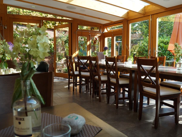 Weincafé De Holter Wienkeller, Wintergarten, Gemeinde Rhauderfehn Helles Weincafé mit Holzmöbeln, langen Tischen und Stühlen, Glasflaschen mit weißen Blumen auf den Tischen, große Fenster mit Blick auf Garten