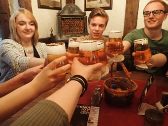Young people laugh and clink glasses of beer at a wooden table in a rustic setting.