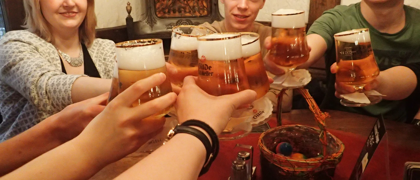 Grüner Jäger Junge Menschen stoßen lachend mit vollen Biergläsern an einem Holztisch in einer rustikalen Umgebung an.