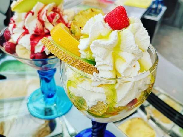 Eiscafé & Pizzeria bei Bruno Rhauderfehn Zwei bunte Eisspezialitäten mit Sahne, Erdbeere, Melone, Ananas und Orangenscheiben in blauen Gläsern vor einer Eistheke