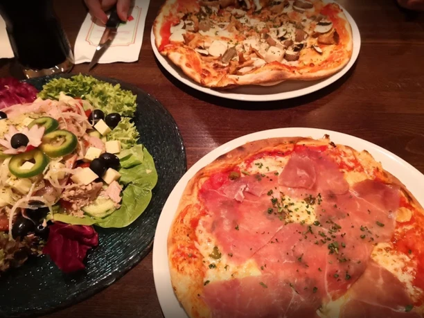 Eiscafé & Pizzeria bei Bruno Rhauderfehn Tisch mit zwei Pizzen, eine mit Schinken und Kräutern, die andere mit Pilzen, und einem Salatteller mit Oliven, Käsewürfeln, Paprika und Salatblättern
