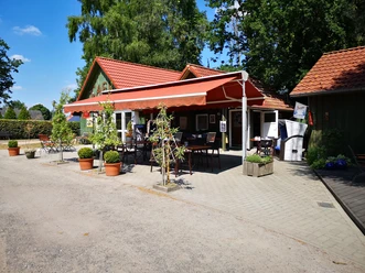 Café am Siel Rhauderfehn Kleines Café mit rotem Sonnenschutz, Tischen und Stühlen im Freien, umgeben von Bäumen und Pflanzen