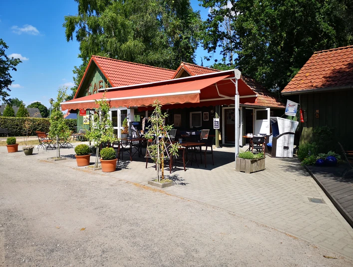 Café am Siel Rhauderfehn Kleines Café mit rotem Sonnenschutz, Tischen und Stühlen im Freien, umgeben von Bäumen und Pflanzen