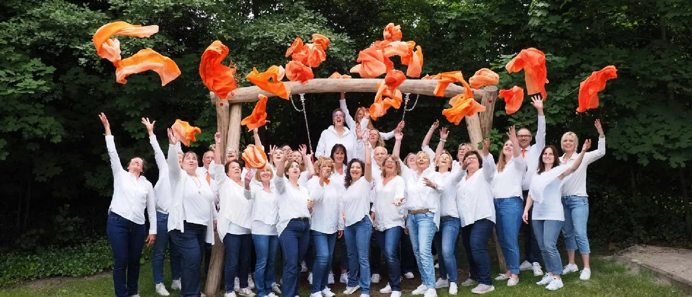 Pressefoto WOF 2024.jpg Ein Chor in weißen Hemden und Jeans wirft im Grünen jubelnd orange Tücher hoch.