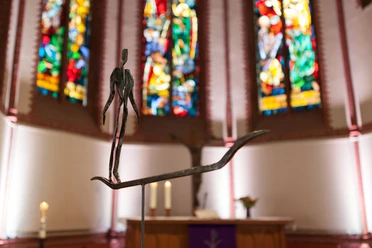 5_Überfahrt_Foto Kai Martin Ulrich_WEB.jpg Schmale Metallfigur auf einem Boot im Altarraum vor bunten Kirchenfenstern.