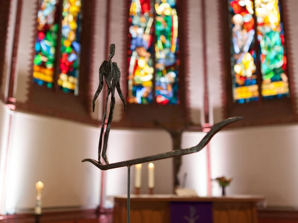 5_Überfahrt_Foto Kai Martin Ulrich_WEB.jpg Schmale Metallfigur auf einem Boot im Altarraum vor bunten Kirchenfenstern.