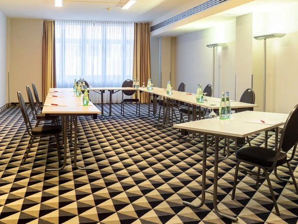 Bright meeting room with U-shaped tables, chairs and drinks on a patterned floor.