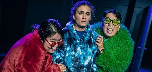 Drei Schauspielerinnen in bunten Pelzjacken und Brillen auf der Bühne, ausdrucksstark beleuchtet