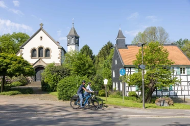 MuseumsLandschaft Abtsküche in Heiligenhaus Zwei Radfahrer_innen passieren eine Kirche und ein historisches Fachwerkhaus an einem sonnigen Tag.