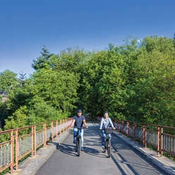 Viadukt Ruhrstraße Süd in Heiligenhaus Zwei Radfahrer_innen auf einer Brücke mit Bäumen und blauem Himmel.