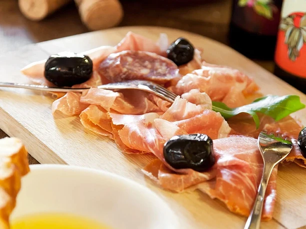 Festa Terrazza Auf einem Brett liegen Prosciutto, Wurstscheiben und schwarze Oliven neben Öl mit Balsamico