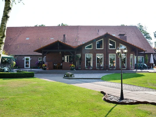 Bauerncafe´Oltmanns Das Bild zeigt das Bauerncafé Oltmanns, ein charmantes Backsteingebäude mit großen, einladenden Fenstern, die aus dem Satteldach hervorragen. Vor dem Gebäude erstreckt sich eine gepflegte Rasenfläche mit einer dekorativen Laterne und Blumenarrangements, die eine idyllische und einladende Atmosphäre schaffen. Im Hintergrund sind Bäume zu sehen, die den ländlichen Charakter der Umgebung betonen.