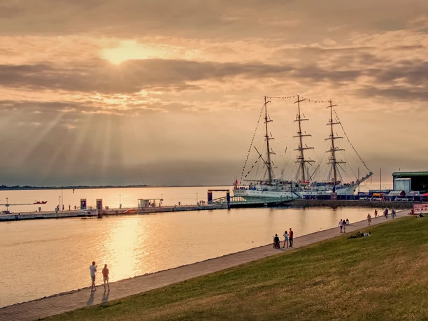 IMG_0472 kopia1 (c) Anna Ściwiarska_Erlebnis Bremerhaven.jpg Spaziergänger auf der Deichpromenade im Gegenlicht. Am Pier liegt ein großes Segelschiff.
