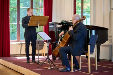 Kurkonzert-Spiegelsaal-Bad-Zwischenahn1.jpg Musiker spielen Geige, Cello und Klavier auf einer Bühne vor großen, hellen Fenstern im Spiegelsaal.