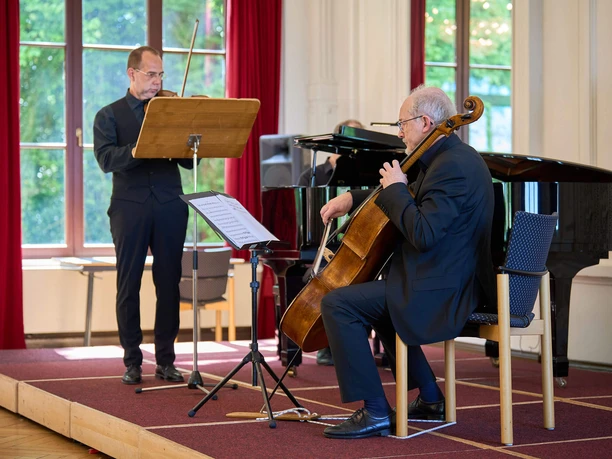 Kurkonzert-Spiegelsaal-Bad-Zwischenahn1.jpg Musiker spielen Geige, Cello und Klavier auf einer Bühne vor großen, hellen Fenstern im Spiegelsaal.