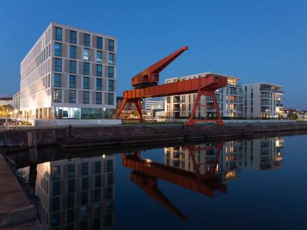 aboutcities_bremerhaven_kran(c)HelmutGross_WEB.jpg Roter historischer Kiesverladekran und moderne Gebäude spiegeln sich abends im Hafenbecken.