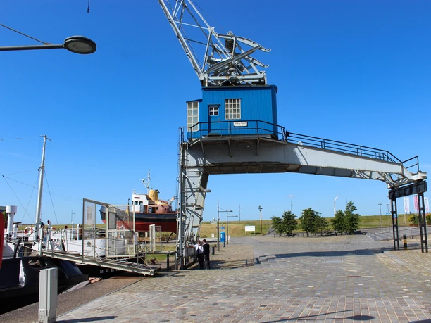 Elektrischer Halbportalkran Havenwelten im Sommer IMG_8644 (c) Tanja Mehl_Erlebnis Bremerhaven_WEB.jpg Ein blauer Halbportalkran am sonnigen Museumshafen überspannt den gepflasterten Weg.