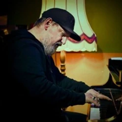 Mann mit Kappe spielt konzentriert Klavier, im Hintergrund steht eine warm leuchtende Stehlampe.