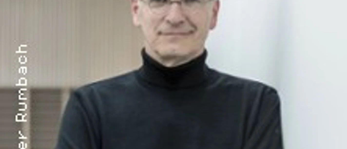 Ein älterer Mann mit Brille im schwarzen Rollkragenpullover lehnt entspannt an einer Wand.