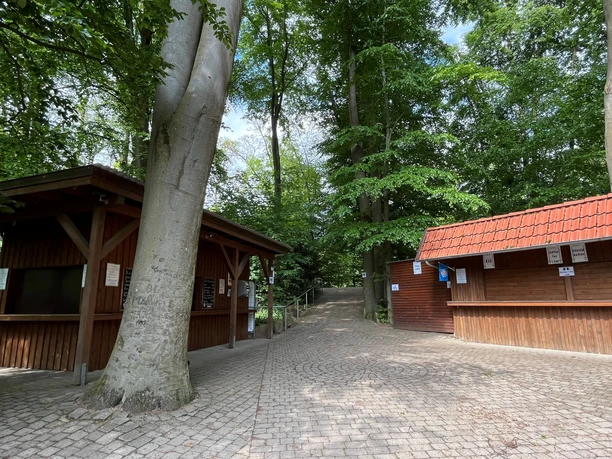 Daverden open-air stage A paved path leads between two wooden kiosks under large trees into the forest.