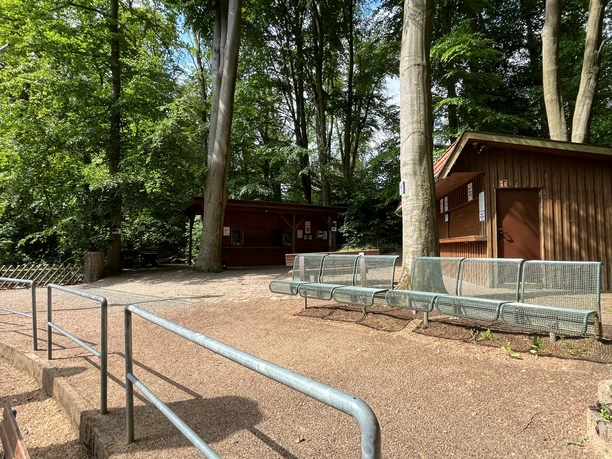 Grüne Metallbänke vor Holzhütten im Schatten hoher Bäume auf der Freilichtbühne.