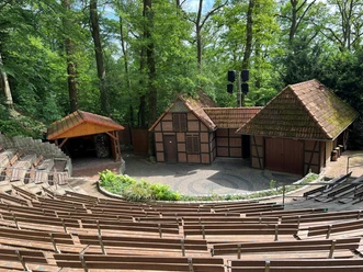 Freilichtbühne Daverden Hölzerne Sitzreihen vor einer Freilichtbühne mit kleinen Fachwerkhäusern im grünen Wald.