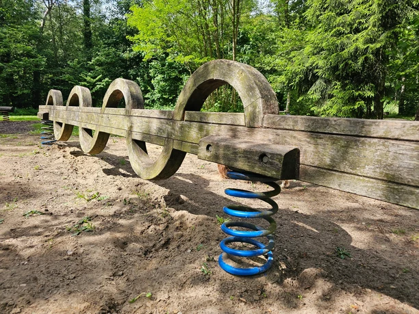 Gesundheitspark Speckenbütterl 20260523_120452 (c) Tanja Mehl_Erlebnis Bremerhaven_WEB.jpg Balanciergerät mit Holzbögen auf blauen Federn auf Sand vor grünen Bäumen im Park.