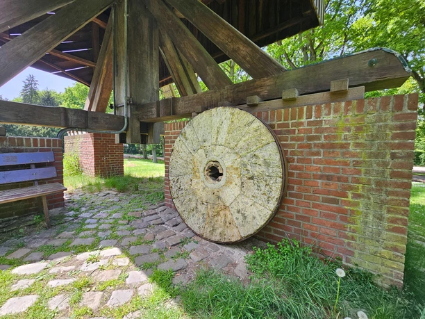 Gesundheitspark Speckenbütterl 20260523_125350 (c) Tanja Mehl_Erlebnis Bremerhaven_WEB.jpg Alter Mühlstein lehnt an einer Backsteinmauer unter dem Holzgerüst der Bockwindmühle.