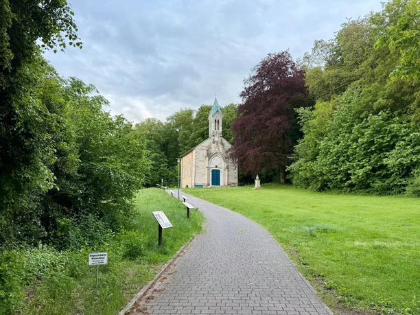 Ein gepflasterter Weg führt zur hellen Friederikenkapelle zwischen grünen Bäumen.