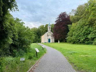 Friederikenkapelle Ein gepflasterter Weg führt zur hellen Friederikenkapelle zwischen grünen Bäumen.