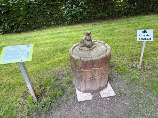 Froschkönig-Holzskulptur auf einem Brunnen mit Infotafel und Schild Küss den Frosch.