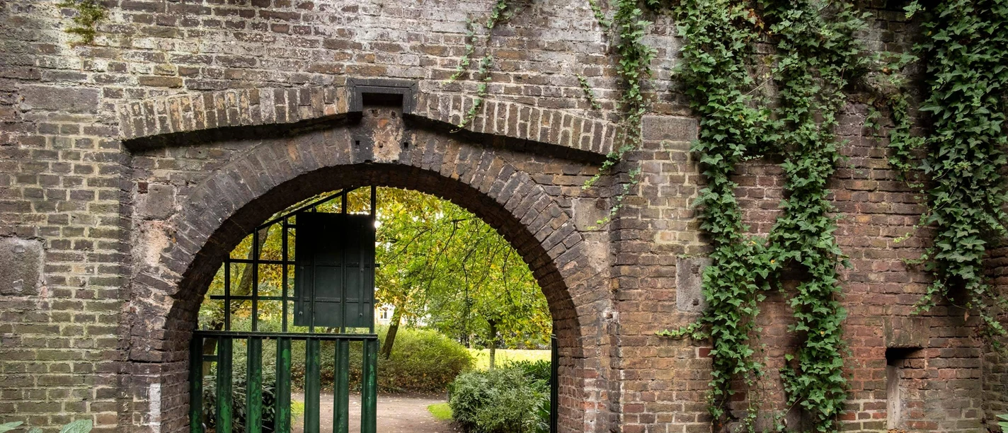 Fort X Alter Backsteinbogen mit geöffnetem grünen Tor und überwachsenen Pflanzen im Parkweg