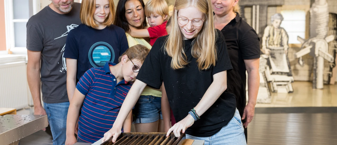 Führung in der Papiermühle Kinder und Erwachsene beobachten ein Mädchen beim Papierschöpfen in einer Museumswerkstatt.