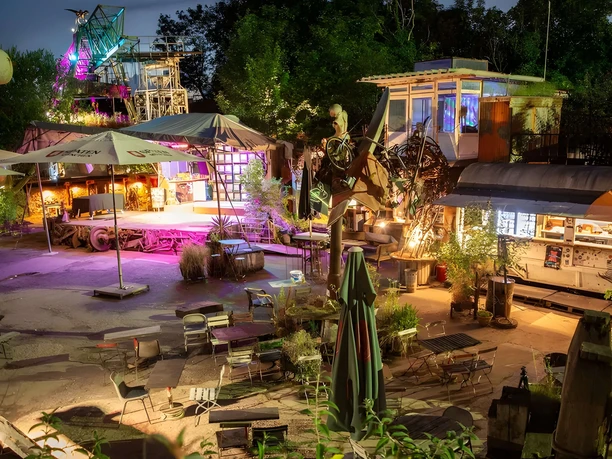 Biergarten Odonien Beleuchteter Außenbereich mit Metallskulpturen, Bühne und Sitzplätzen in kreativem Ambiente