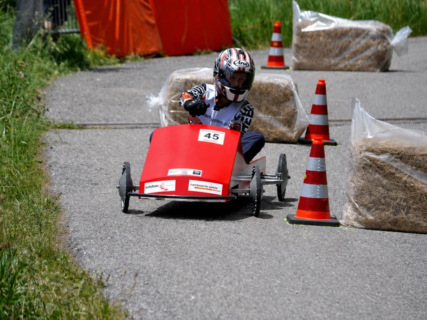 Header_optimized.png A racer steering a small soapbox car through cones and hay bales on a paved track