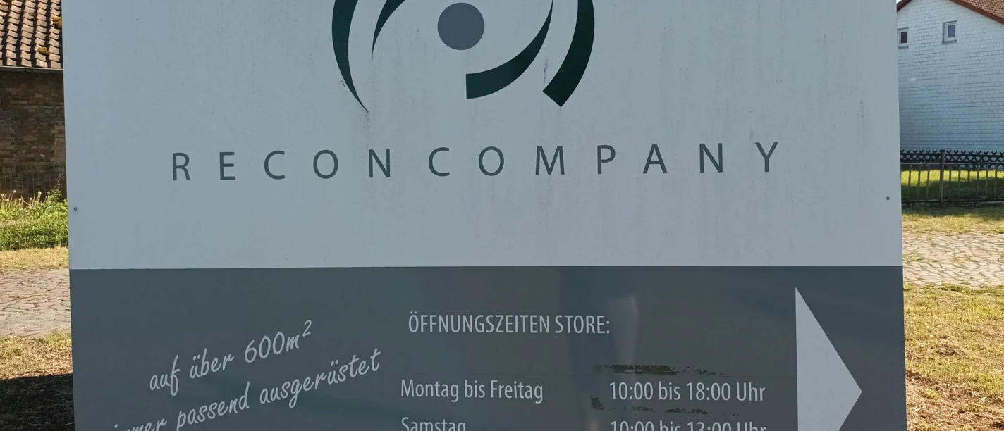 Recon Company_Salzgitter_HD_16zu9.jpg Firmenschild der Recon Company mit Logo, Öffnungszeiten und einem Pfeil nach rechts.