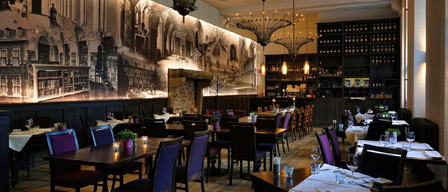 Kloosterbrasserie de Kanunnik Innenraum des Restaurants mit mehreren Sitzgruppen und einem hohen Wandregal mit verschiedenen Weinflaschen. Von der Decke hängen mehrere Lampen herab.