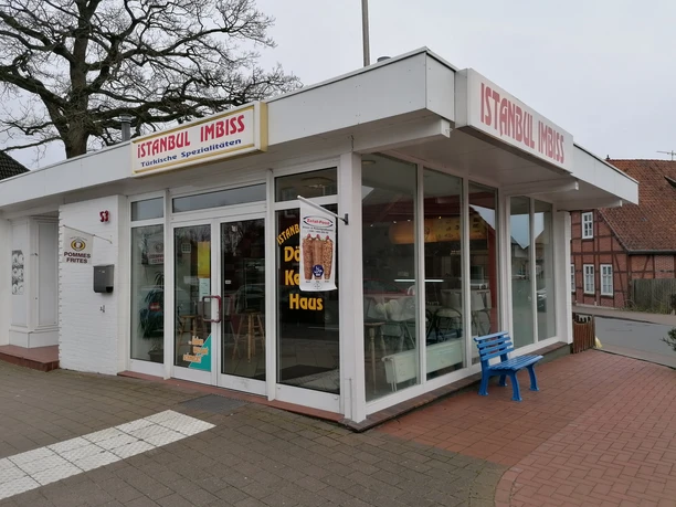 Außenansicht Weißes Flachdachgebäude mit großen Glasfenstern, roter Aufschrift Istanbul Imbiss und blauer Bank.