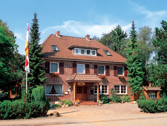 Landgasthaus Tödter Zweigeschossiges Landgasthaus aus rotem Klinker mit Walmdach und weißen Fensterrahmen, umgeben von hohen Nadelbäumen und gepflegtem Grün. Über dem hölzernen Eingangsbereich mit Vordach hängt ein Schild mit dem Namen des Hauses, davor blühen bunte Blumen, eine Fahne weht im Vorgarten.