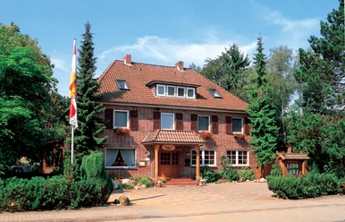 Landgasthaus Tödter Zweigeschossiges Landgasthaus aus rotem Klinker mit Walmdach und weißen Fensterrahmen, umgeben von hohen Nadelbäumen und gepflegtem Grün. Über dem hölzernen Eingangsbereich mit Vordach hängt ein Schild mit dem Namen des Hauses, davor blühen bunte Blumen, eine Fahne weht im Vorgarten.