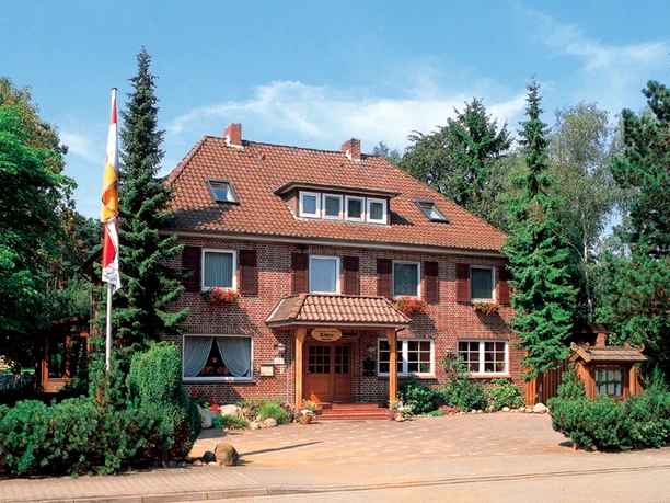 Landgasthaus Tödter Zweigeschossiges Landgasthaus aus rotem Klinker mit Walmdach und weißen Fensterrahmen, umgeben von hohen Nadelbäumen und gepflegtem Grün. Über dem hölzernen Eingangsbereich mit Vordach hängt ein Schild mit dem Namen des Hauses, davor blühen bunte Blumen, eine Fahne weht im Vorgarten.