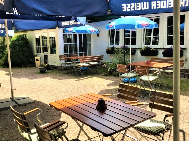 Außenbereich Sonnige Außenterrasse mit blauen Schirmen, Holztischen und Stühlen vor einer weißen Hausfassade.