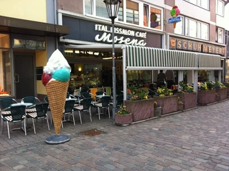eiscafe-mosena-aussen Großes Eiswaffel-Dekorationselement vor Eiscafé Mosena, umgeben von Außensitzplätzen und Blumenkästen.