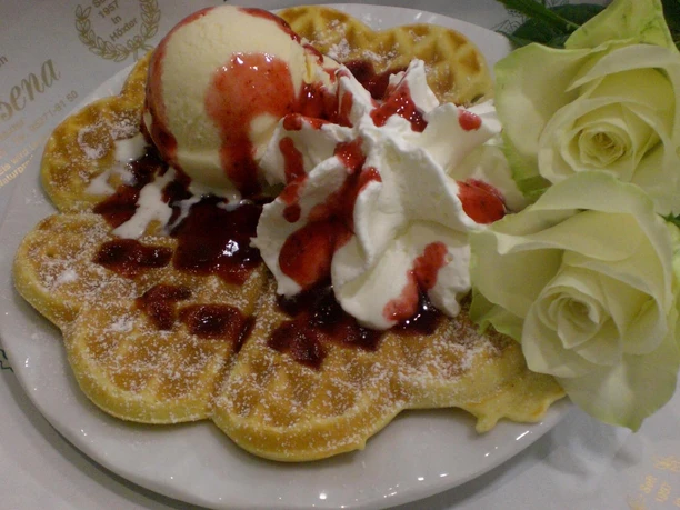 Waffel mit Vanilleeiskugel, fruchtiger Erdbeersauce, Sahnehäubchen und gelben Rosen auf einem Teller.
