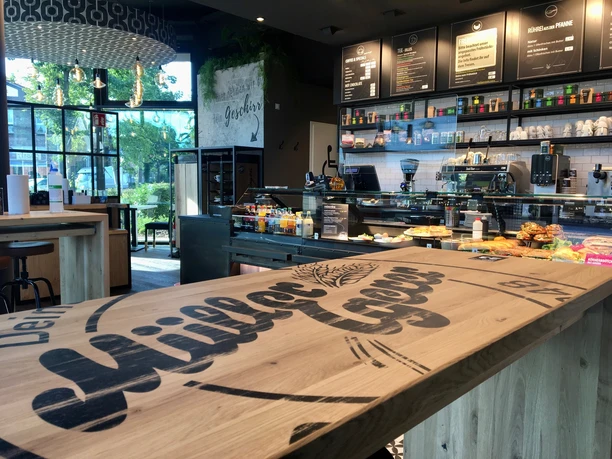 Müller & Egerer Das Bild zeigt das Interieur einer modernen Bäckerei mit einem großen Holzverkaufstresen von "Müller & Egerer". Im Hintergrund sind Regale mit Produkten und eine Kaffeemaschine zu sehen. Der Raum ist hell, mit großen Fenstern und stilvoller Beleuchtung.