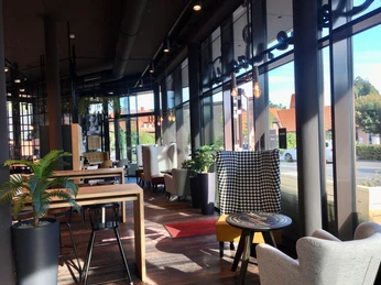 Müller & Egerer Ein gemütliches Café mit hohen Fenstern, stilvollen Polstermöbeln und viel natürlichem Licht. Pflanzen stehen in modernen Töpfen und Holzmöbel verleihen dem Raum Wärme.